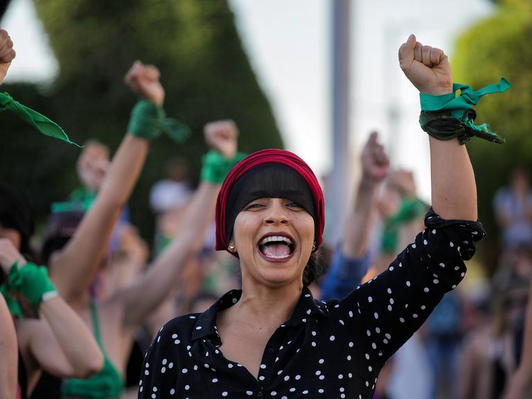 Im Vordergrund einer Gruppe von Frauen steht eine Frau mit erhobener Faust. Sie trägt am Arm schwarze und grüne Bänder.