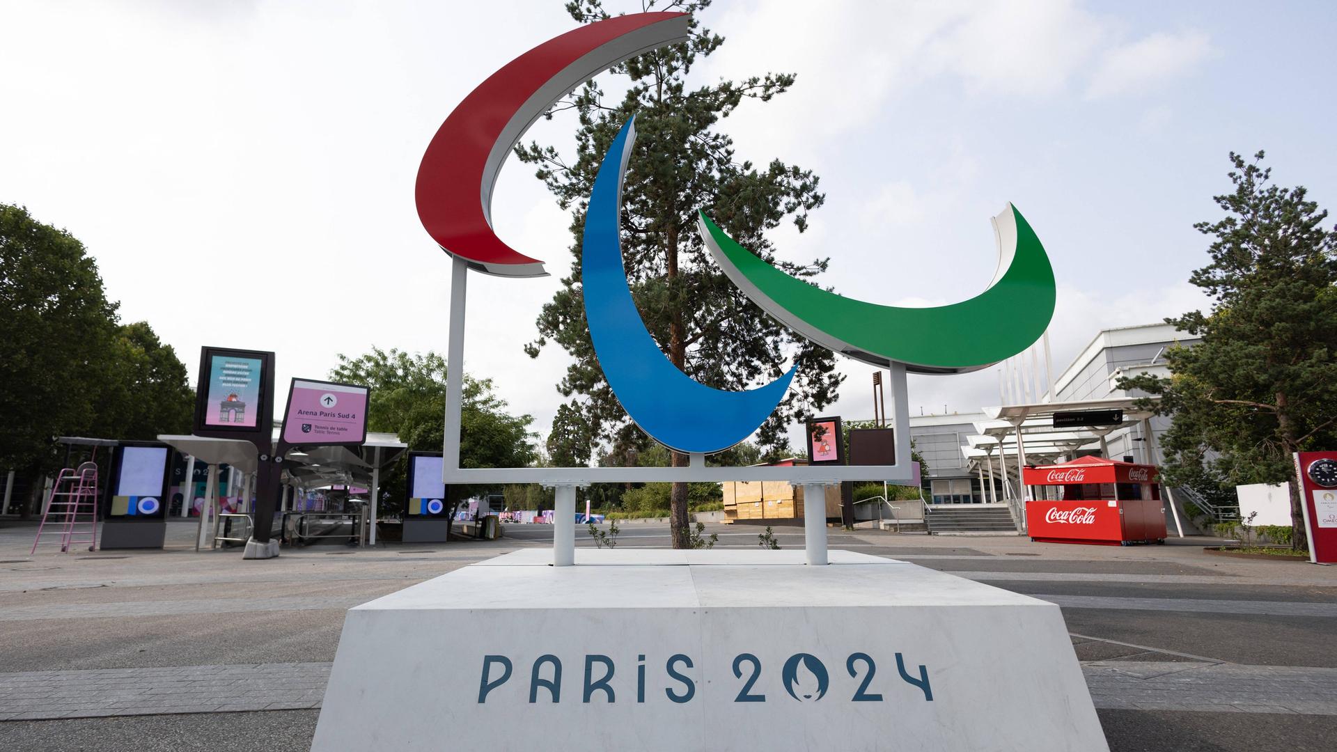Auf einem Platz steht eine Skulptur mit dem rot-blau-grünen Paralympics-Symbol. Darunter steht: Paris 2024. Auf einem Platz steht eine Skulptur mit dem rot-blau-grünen Paralympics-Symbol. Darunter steht: Paris 2024.