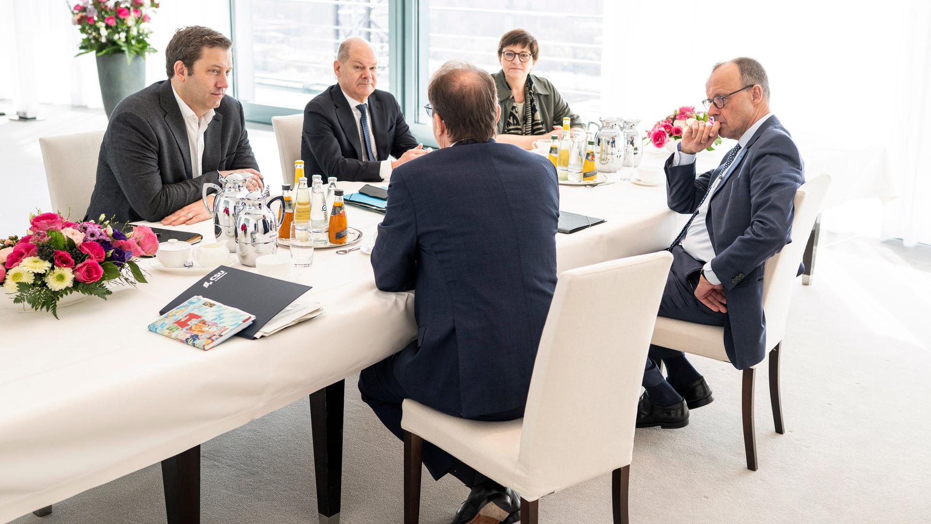 Die vier sitzen in einem Raum an einem weißen Tisch auf weißen Stühlen. 