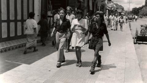 Drei junge indonesische Frauen laufen eine Straße entlang. Die äußeren tragen militärische Kleidung, die in der Mitte ein weißes Kleid.