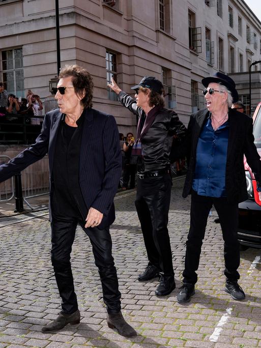 Ronnie Wood, Mick Jagger und Keith Richards stehen auf einer Straße vor einem Taxi und winken Journalisten.