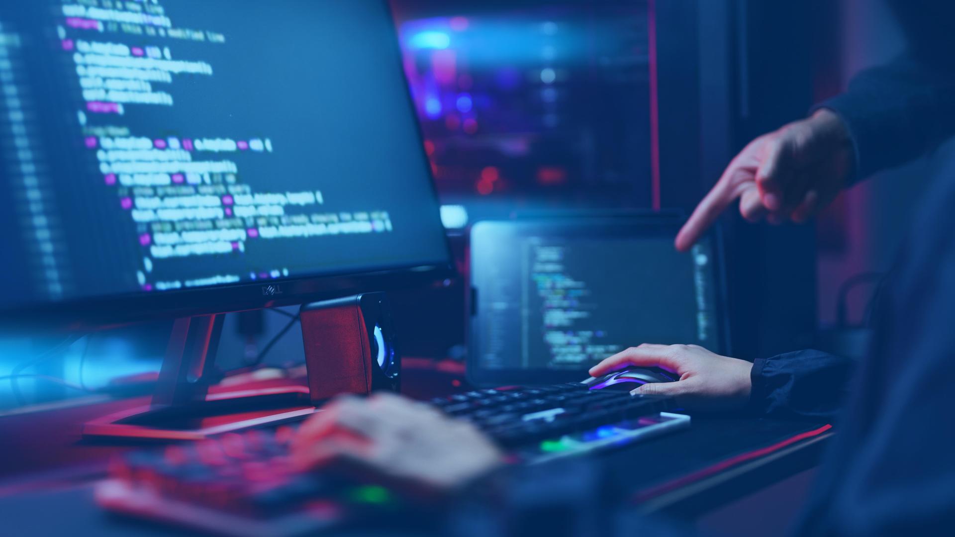 Auf zwei Monitoren sind Programmiercode und technische Oberflächen zu sehen. Eine Person sitzt an der Tastatur, eine weitere Hand zeigt auf den Bildschirm. 
