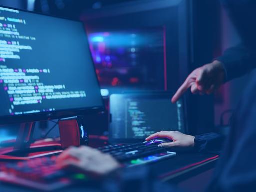 Auf zwei Monitoren sind Programmiercode und technische Oberflächen zu sehen. Eine Person sitzt an der Tastatur, eine weitere Hand zeigt auf den Bildschirm. 