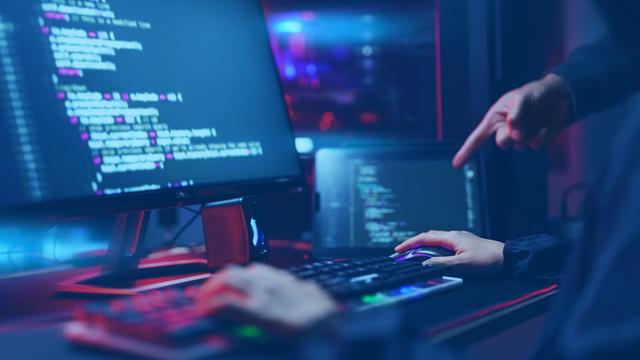 Auf zwei Monitoren sind Programmiercode und technische Oberflächen zu sehen. Eine Person sitzt an der Tastatur, eine weitere Hand zeigt auf den Bildschirm. 