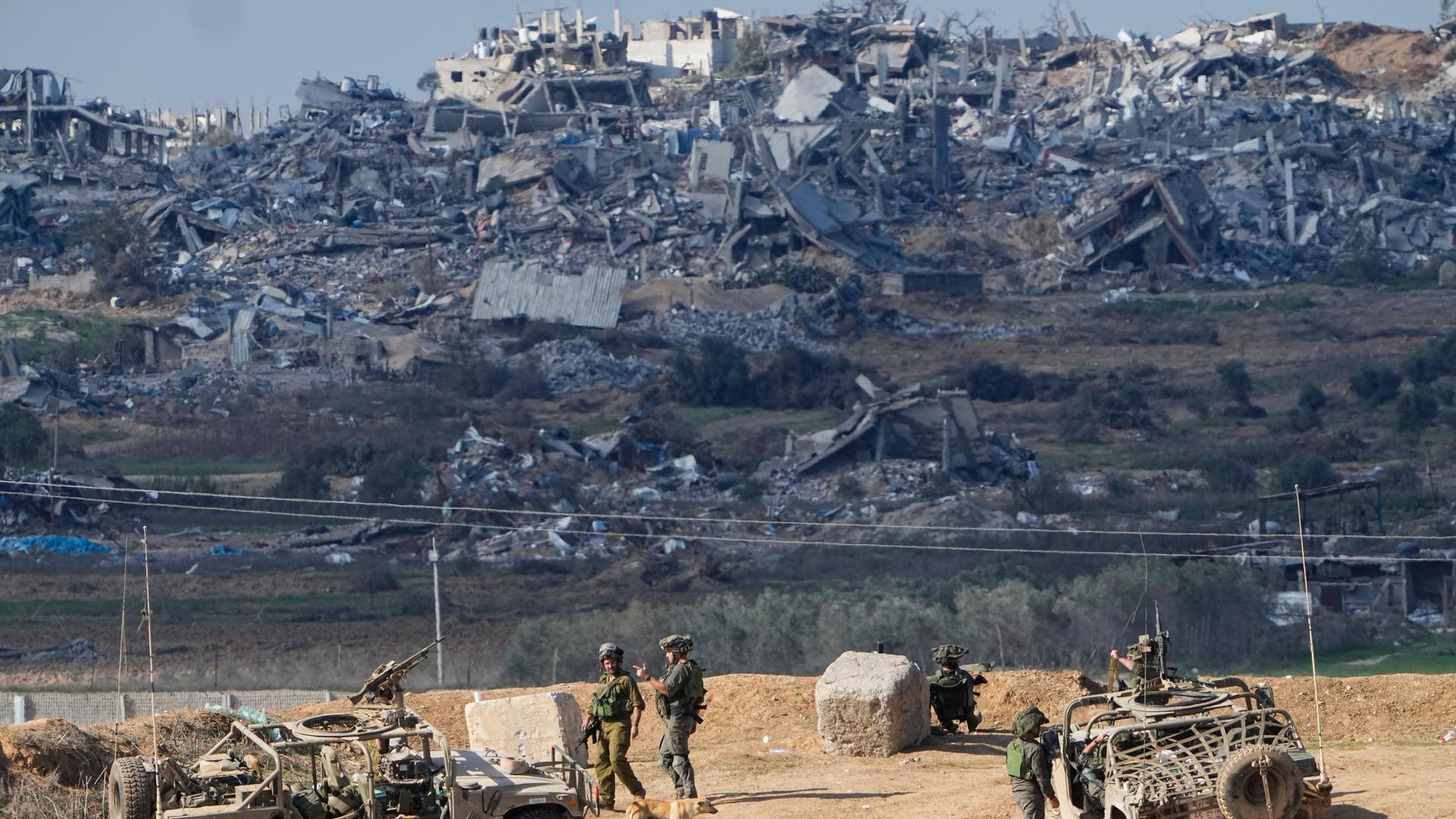 Zwei Jeeps mit Maschinengewehren und mehrere israelische Soldaten stehen auf einem Hügel. Im Hintergrund sind zerstörte Häuser zu sehen.
