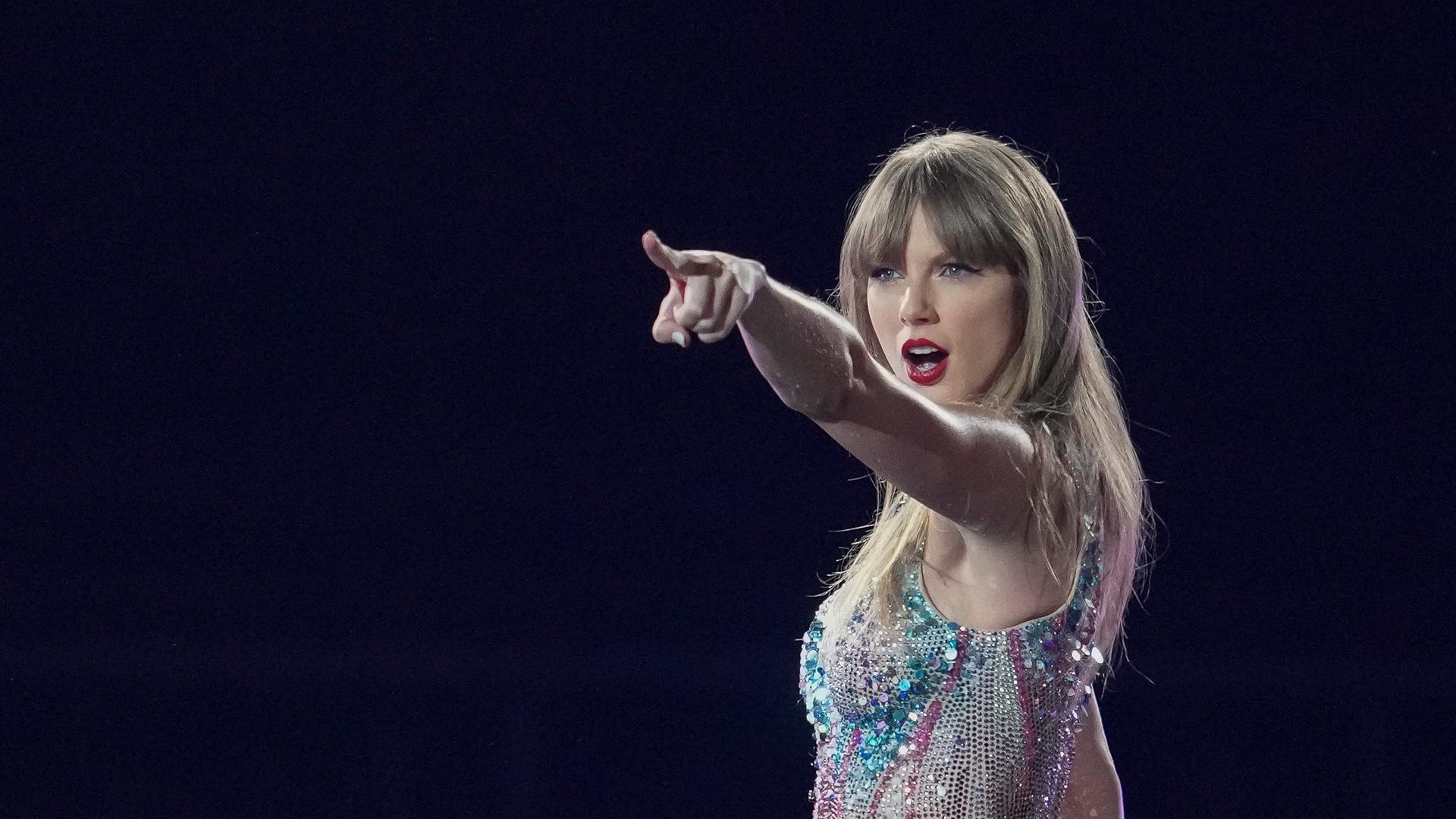 Die US-Sängerin Taylor Swift steht auf einer dunklen Bühne, trägt einen Glitzerbody und zeigt mit dem Finger ins Publikum.