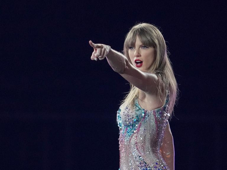 Die US-Sängerin Taylor Swift steht auf einer dunklen Bühne, trägt einen Glitzerbody und zeigt mit dem Finger ins Publikum.
