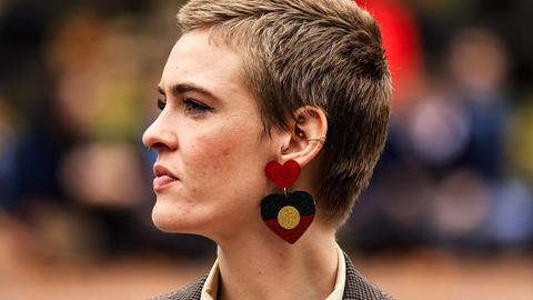 Eine junge Frau mit kurzen Haaren und mit Ohrringen in Herzform, die die Flagge der Aborigines zeigt, ist von der Seite zu sehen.