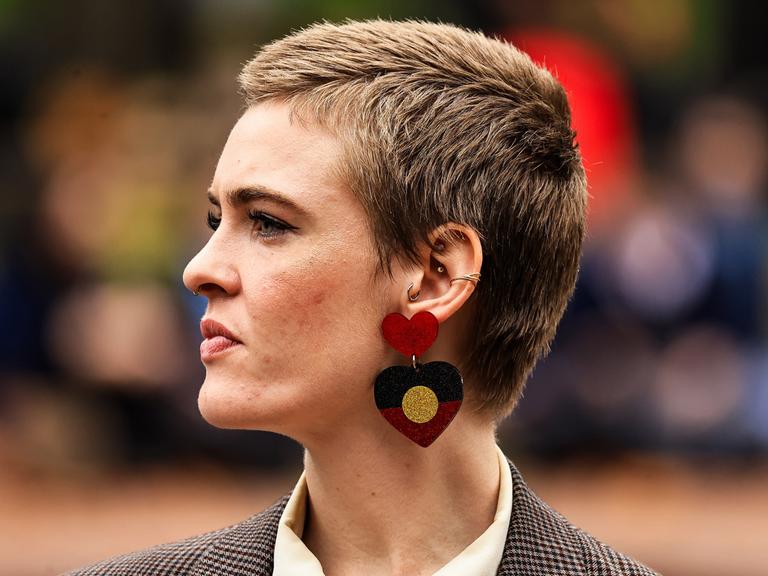 Eine junge Frau mit kurzen Haaren und mit Ohrringen in Herzform, die die Flagge der Aborigines zeigt, ist von der Seite zu sehen.