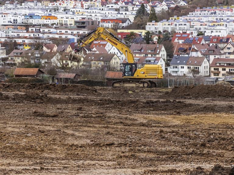 Ein Bagger steht auf Ackerland. Im Hintergrund sieht man mehrere Häuser. Ein Bagger steht auf Ackerland. Im Hintergrund sieht man mehrere Häuser.