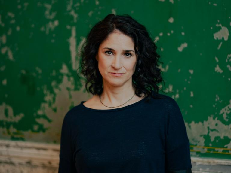 Porträtfoto der deutsch-ägyptischen Journalistin Annabel Wahba. Sie hat dunkles, langes Haar, trägt einen dunklen Pullover und steht vor einer grüngestrichenen Wand, von der die Farbe abblättert.