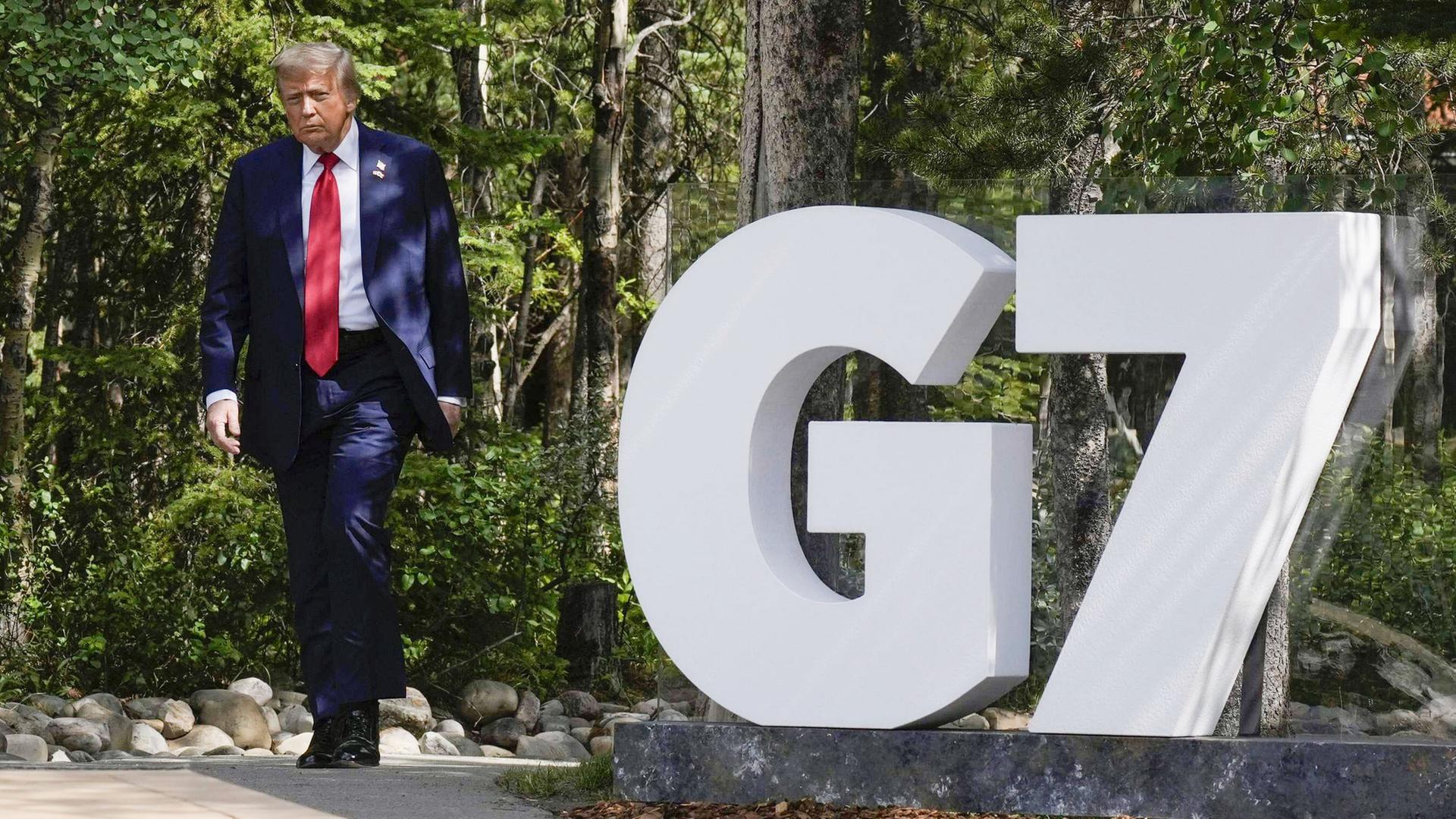 Ein Mann mit blauen Anzug läuft einen Weg entlang. Es ist US-Präsident Donald Trump. Neben ihm ist ein weißes G und eine Sieben. 