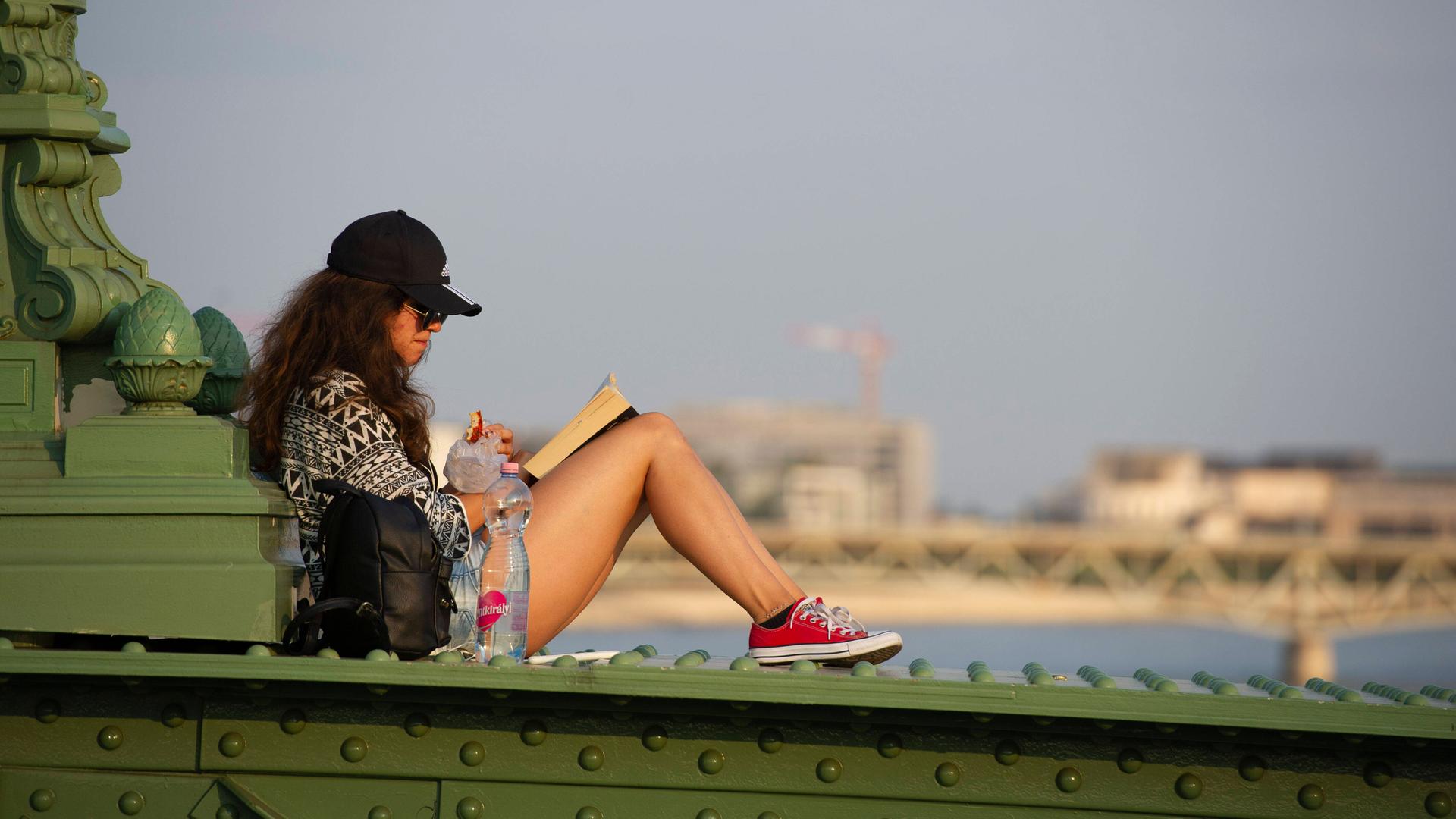 Eine junge Frau sitzt auf der Brüstung der Freiheitsbrücke in Budapest und liest ein Buch.