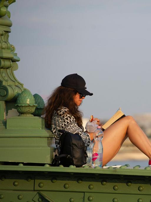 Eine junge Frau sitzt auf der Brüstung der Freiheitsbrücke in Budapest und liest ein Buch.