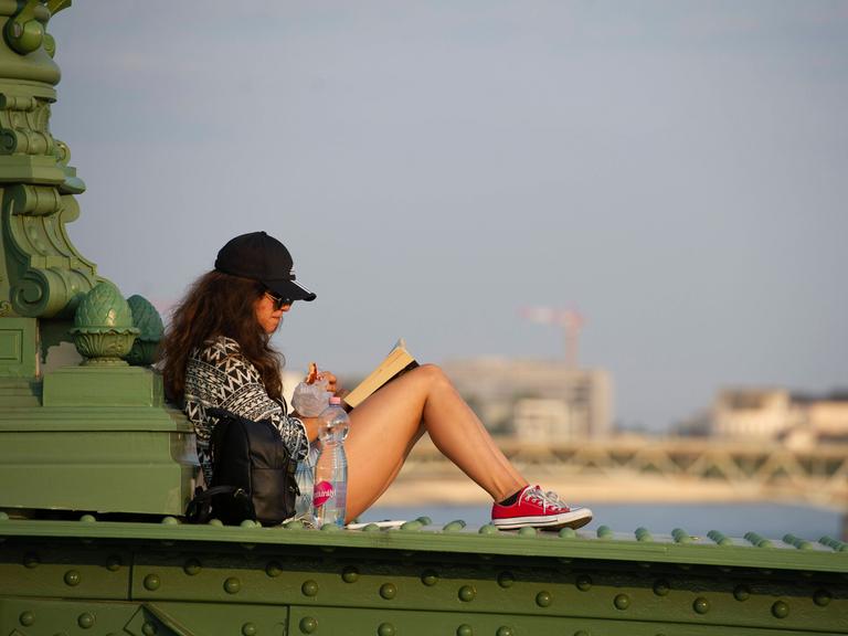 Eine junge Frau sitzt auf der Brüstung der Freiheitsbrücke in Budapest und liest ein Buch.