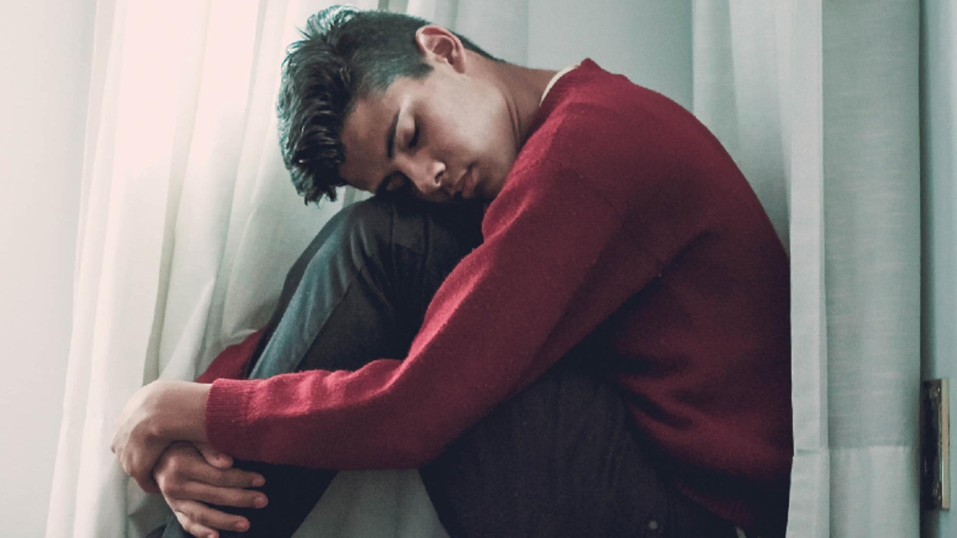 Ein Junge sitzt an einem Fenster hinter einer Gardine. 