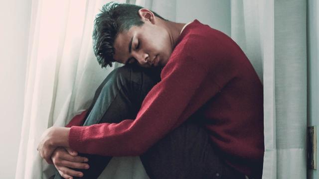 Ein Junge sitzt an einem Fenster hinter einer Gardine. 