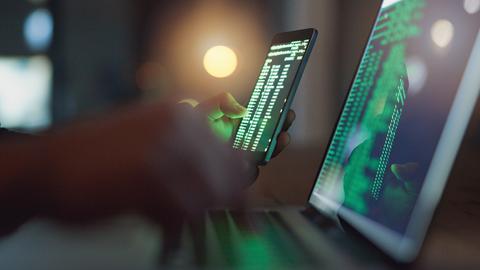 Eine Person sitzt im Halbdunkel mit einem Smartphone vor einem Laptop. Beide Bildschirme zeigen Code.