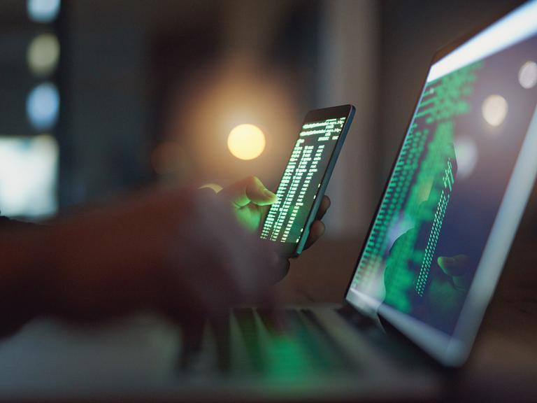 Eine Person sitzt im Halbdunkel mit einem Smartphone vor einem Laptop. Beide Bildschirme zeigen Code.