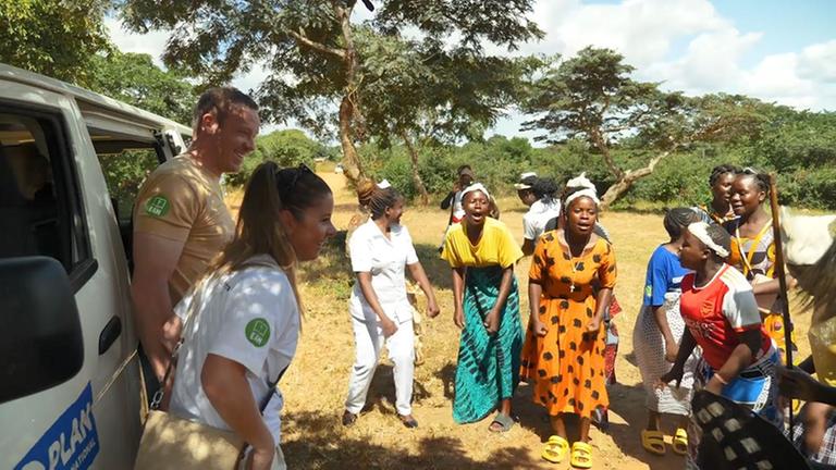 Felix und Lisa Loch steigen aus einem Transporter von Plan International aus und werden von singenden und tanzenden Frauen in Malawi empfangen.