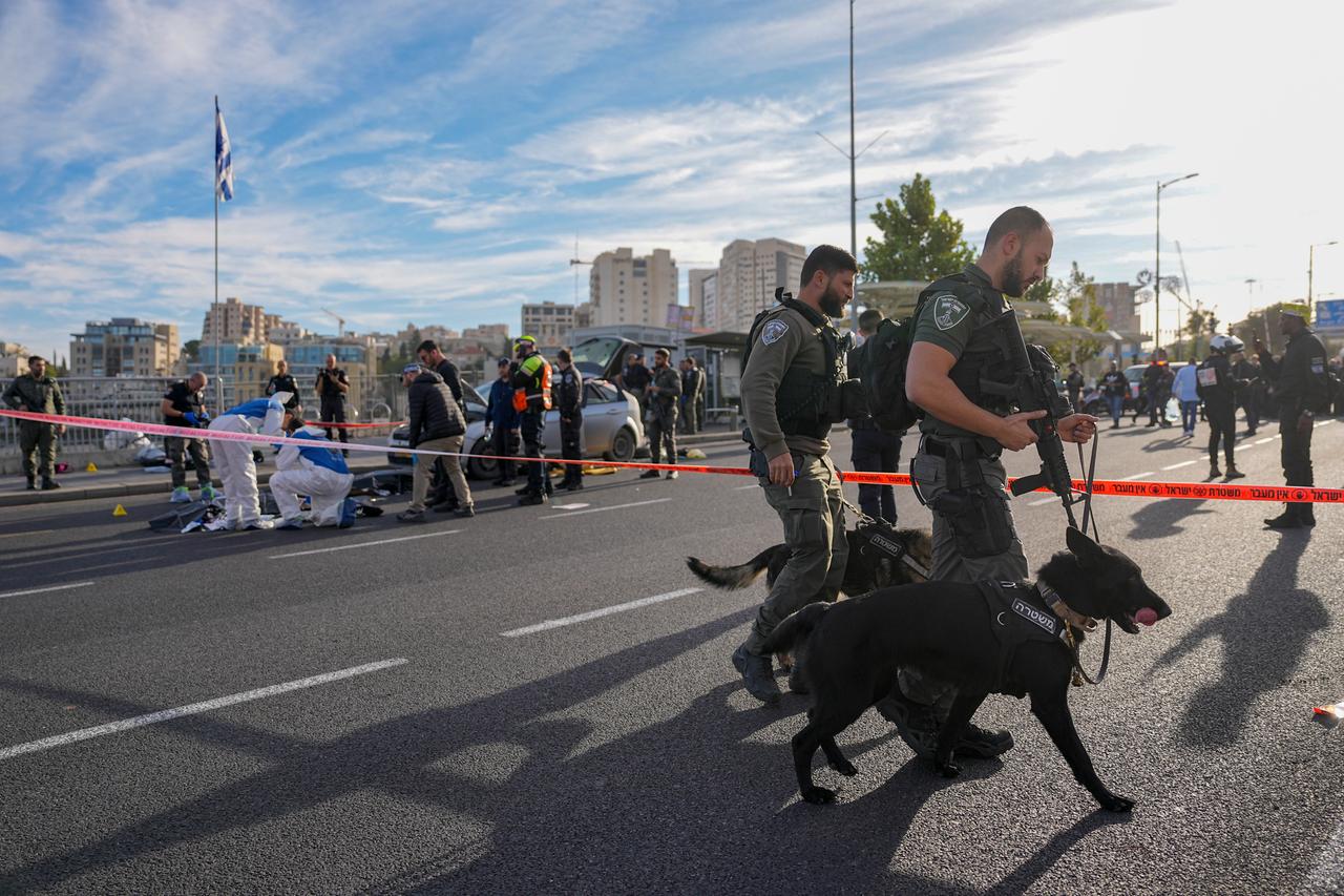 Zwei Soldaten mit Langwaffen und Hunden hinter einer Polizeiabsperrung auf einer Straße.