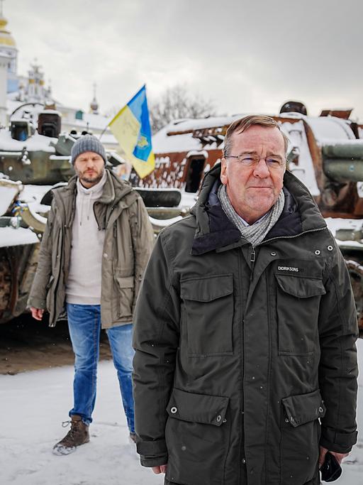Boris Pistorius steht in einer olivgrünen Jacke vor zerstörten russischen Panzern in Kiew. Eine blau-gelbe Fahne ist im Hintergrund zu sehen.