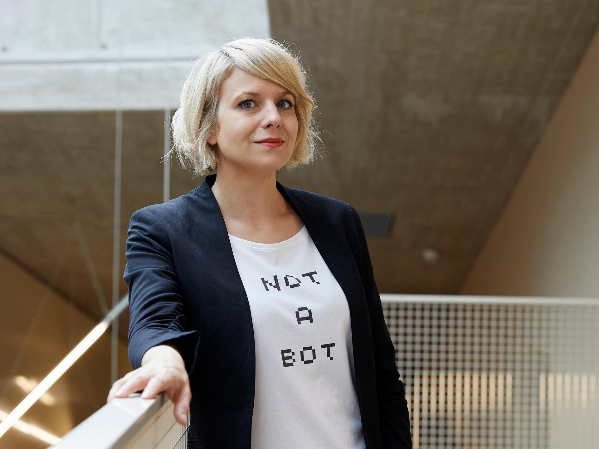 Professorin Martina Mara an der Johannes Kepler Univesität Linz. Sie steht an ein Geländer gelehnt. Mara trägt eine blaue Jeans, eine schwarze Jacke und ein weißes T-Shirt, auf dem „Not a Bot“ steht.
