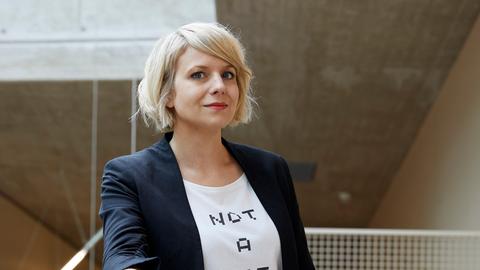 Professorin Martina Mara an der Johannes Kepler Univesität Linz. Sie steht an ein Geländer gelehnt. Mara trägt eine blaue Jeans, eine schwarze Jacke und ein weißes T-Shirt, auf dem „Not a Bot“ steht.