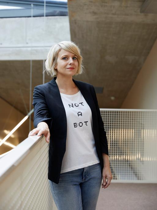 Professorin Martina Mara an der Johannes Kepler Univesität Linz. Sie steht an ein Geländer gelehnt. Mara trägt eine blaue Jeans, eine schwarze Jacke und ein weißes T-Shirt, auf dem „Not a Bot“ steht.