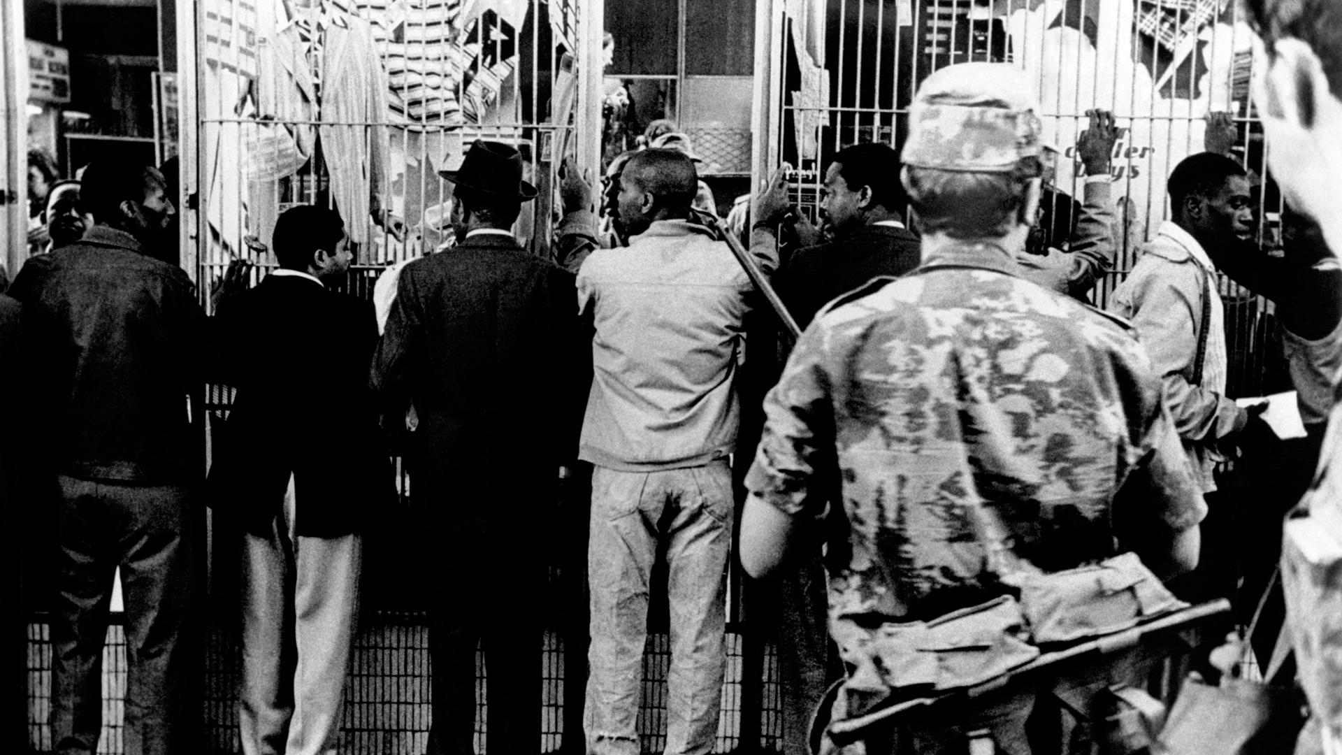 Historisches Schwarzweißfoto Schwarzer Männer, die aufgereiht an einer Wand stehen und von einem bewaffneten weißen Polizisten bewacht werden. Historisches Schwarzweißfoto Schwarzer Männer, die aufgereiht an einer Wand stehen und von einem bewaffneten weißen Polizisten bewacht werden.