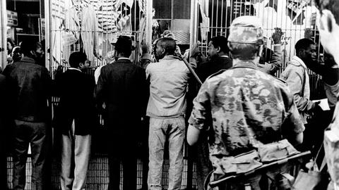 Historisches Schwarzweißfoto Schwarzer Männer, die aufgereiht an einer Wand stehen und von einem bewaffneten weißen Polizisten bewacht werden.