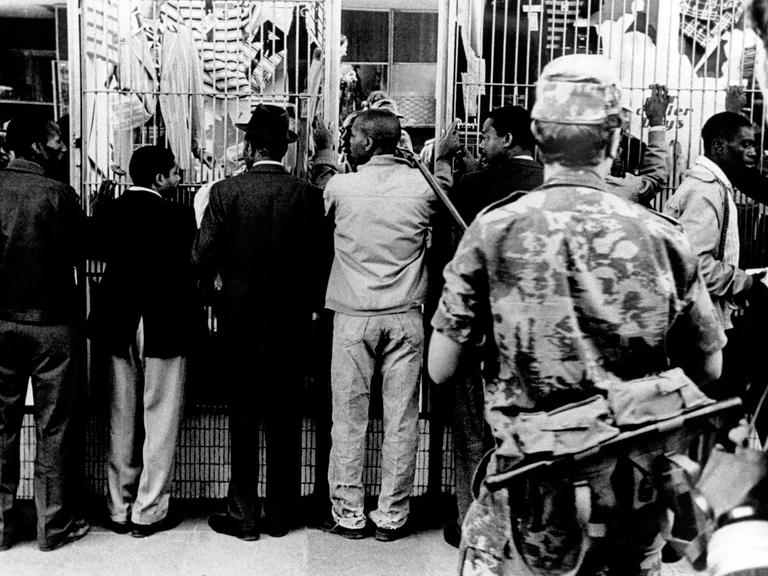 Historisches Schwarzweißfoto Schwarzer Männer, die aufgereiht an einer Wand stehen und von einem bewaffneten weißen Polizisten bewacht werden. Historisches Schwarzweißfoto Schwarzer Männer, die aufgereiht an einer Wand stehen und von einem bewaffneten weißen Polizisten bewacht werden.
