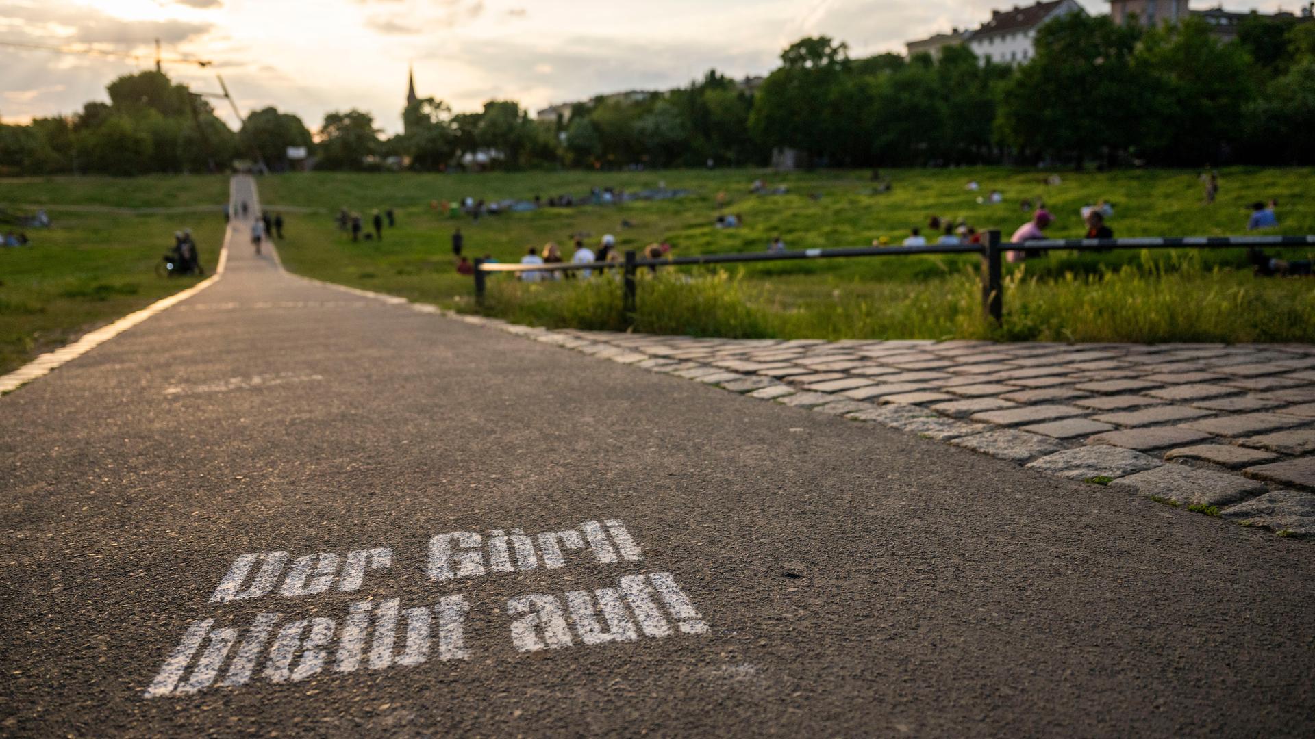 Besucher sitzen bei Sonnenuntergang im Görlitzer Park. Auf dem Asphalt ist "Der Görli bleibt auf!" gesprüht. Besucher sitzen bei Sonnenuntergang im Görlitzer Park. Auf dem Asphalt ist "Der Görli bleibt auf!" gesprüht.