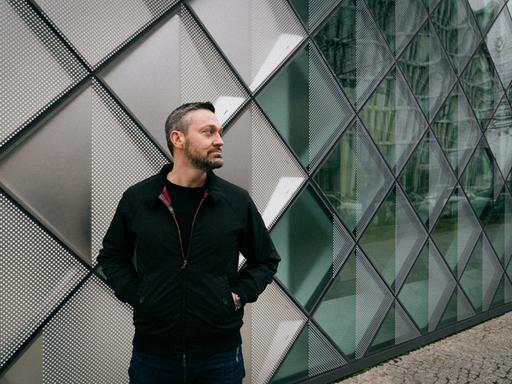 Fritz Kalkbrenner steht mit kurzem Haar und dunkler Jacke vor einer geometrisch gegliederten Wand.