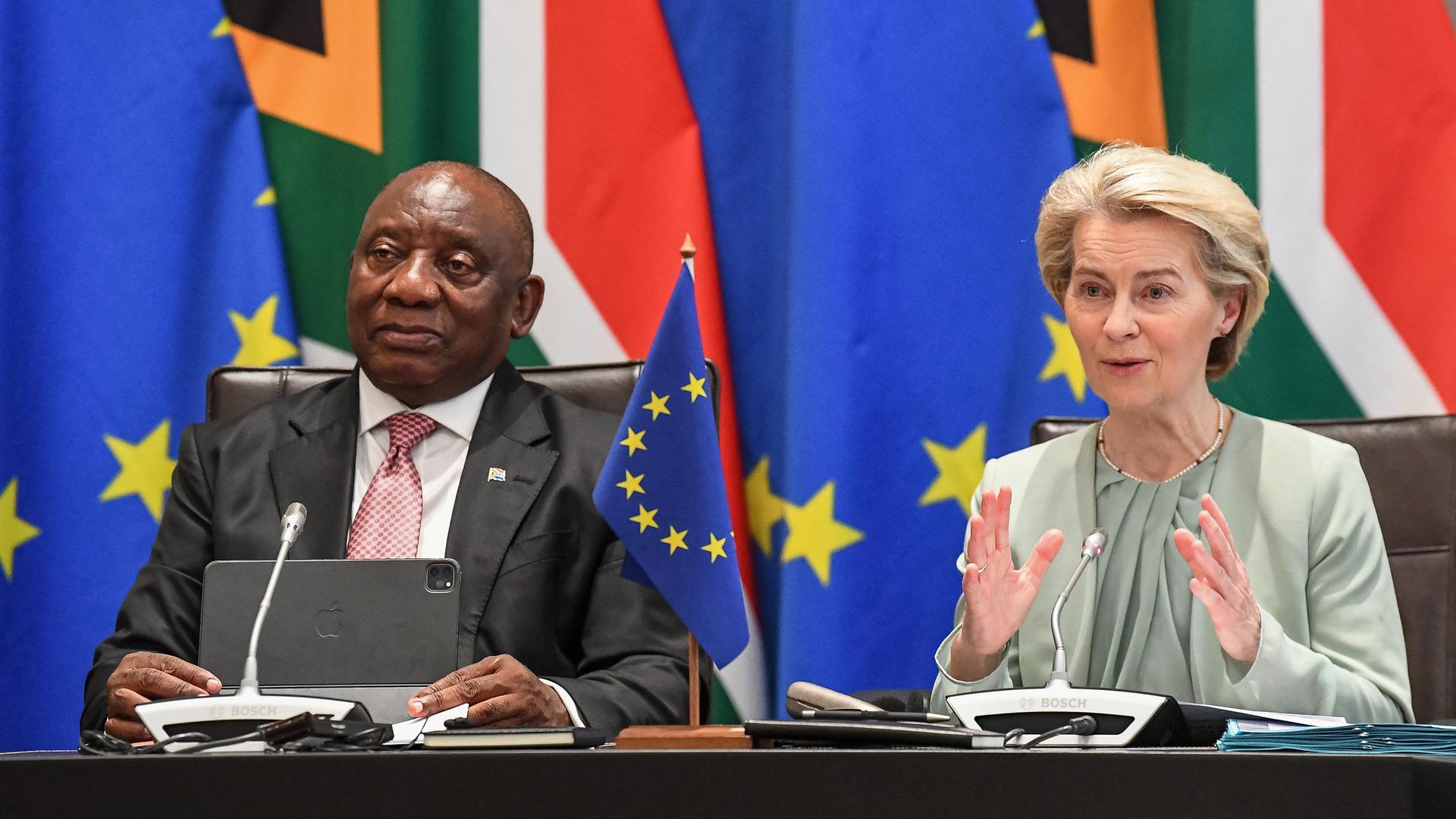 Südafrikas Präsident Ramaphosa (l.) und EU-Kommissionspräsidentin von der Leyen sitzen nebeneinander an einem Tisch. Von der Leyen spricht in ein Mikrophon. Hinter den beiden Politikern sind die Flaggen der EU und Südafrikas zu erkennen. 