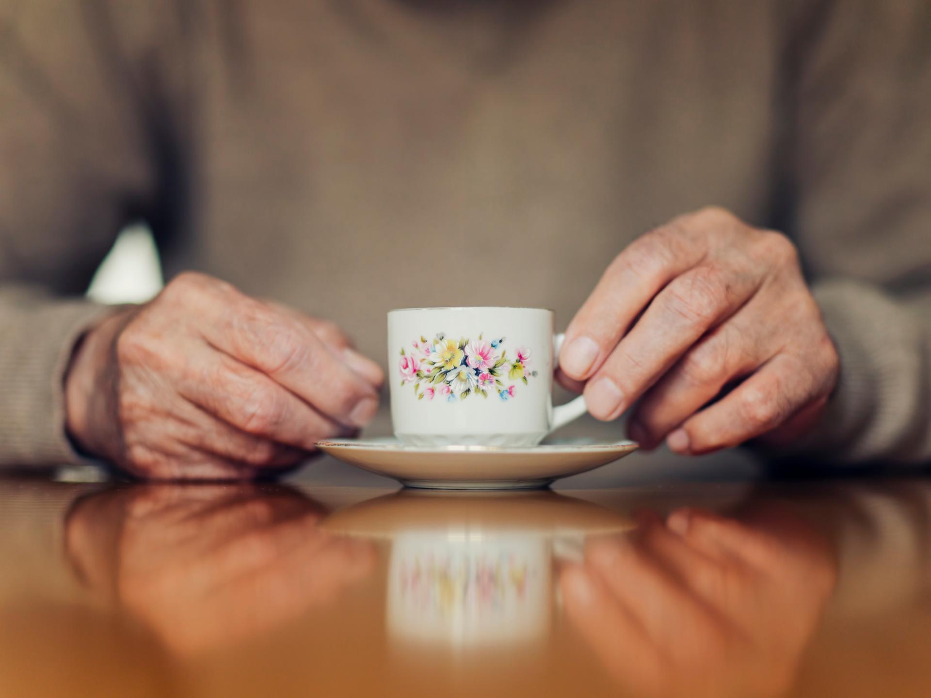 Die Hände eines älteren Menschen halten eine Blümchentasse.