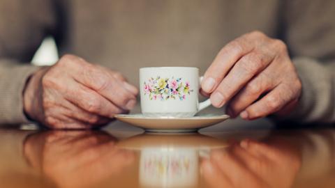 Die Hände eines älteren Menschen halten eine Blümchentasse.