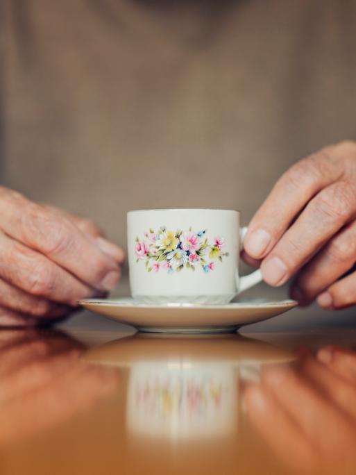 Die Hände eines älteren Menschen halten eine Blümchentasse.