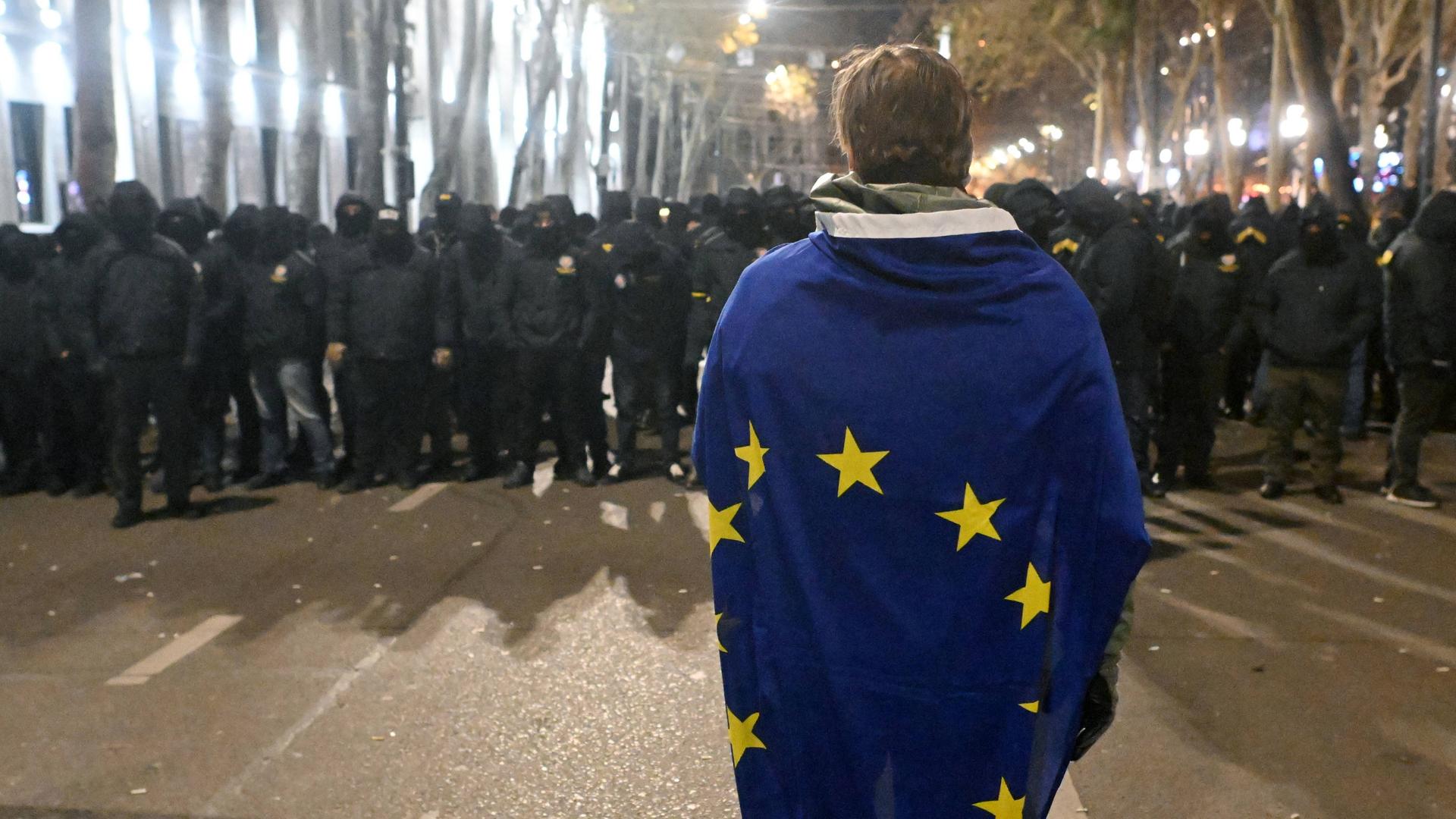 In Tiflis steht eine Person eingehüllt in die EU-Flagge vor einer Menschengruppe, die komplett in schwarz gekleidet ist.