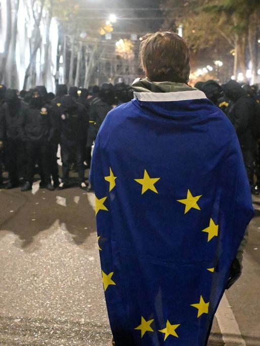 In Tiflis steht eine Person eingehüllt in die EU-Flagge vor einer Menschengruppe, die komplett in schwarz gekleidet ist.