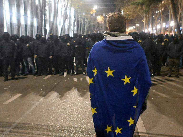 In Tiflis steht eine Person eingehüllt in die EU-Flagge vor einer Menschengruppe, die komplett in schwarz gekleidet ist.