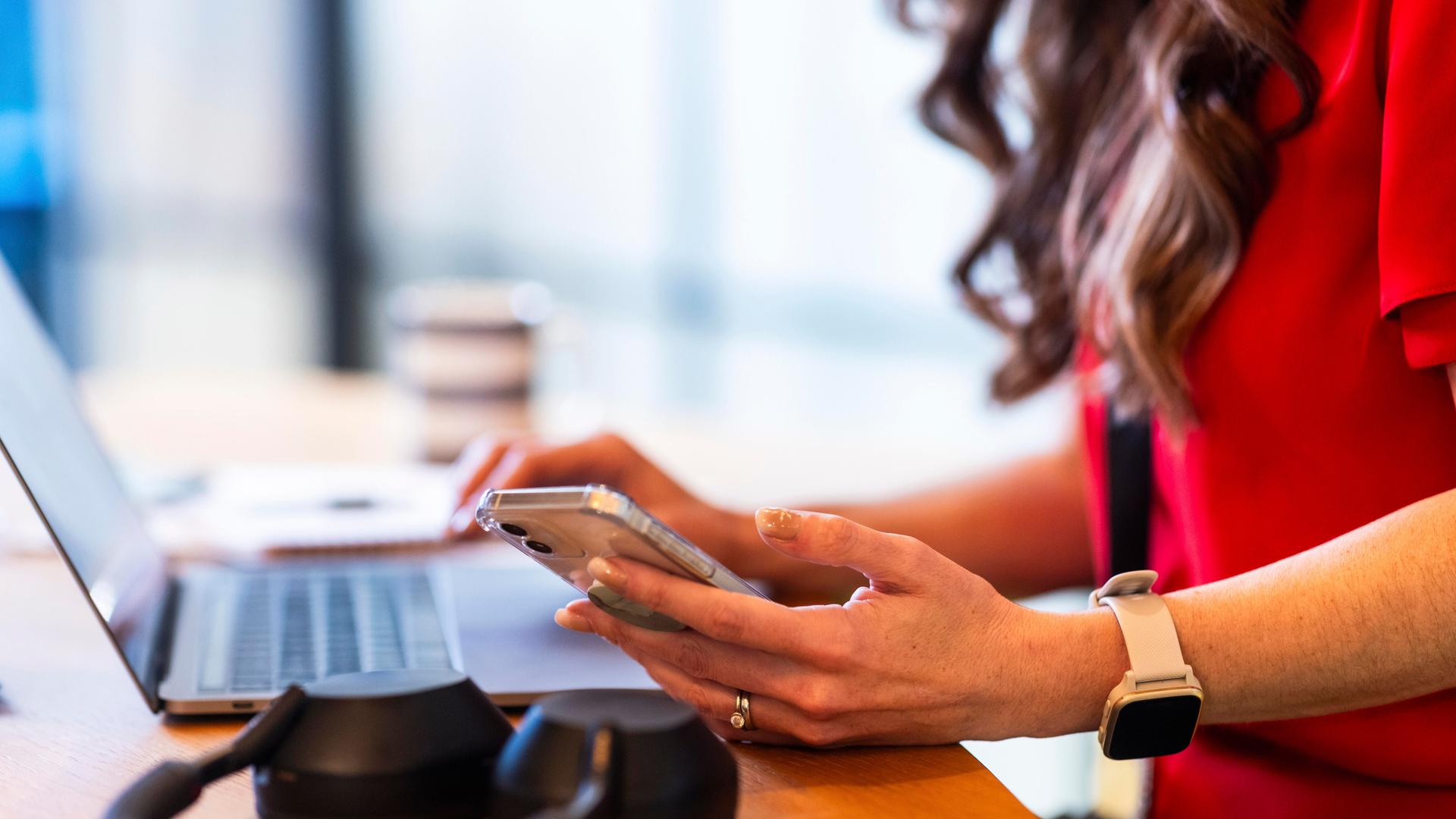 Ein Frau arbeitet am Notebook und hält ein Smartphone in der Hand. Ihre langen braunen Haare fallen ins Bild.