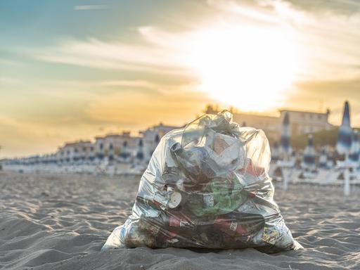 Ein gefüllter Müllsack mit Plastikabfällen liegt im Sand an einem Strand.