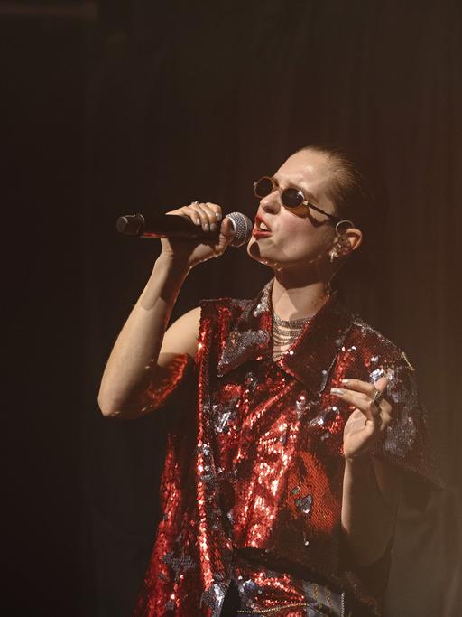 Eine Frau im roten Glitzer-Oberteil steht auf der Bühne mit einem Mikrophon und singt.