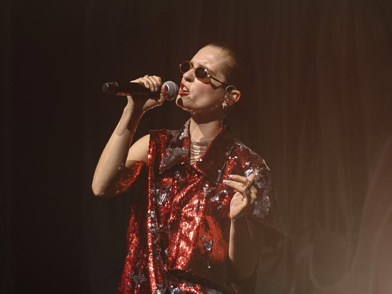 Eine Frau im roten Glitzer-Oberteil steht auf der Bühne mit einem Mikrophon und singt.