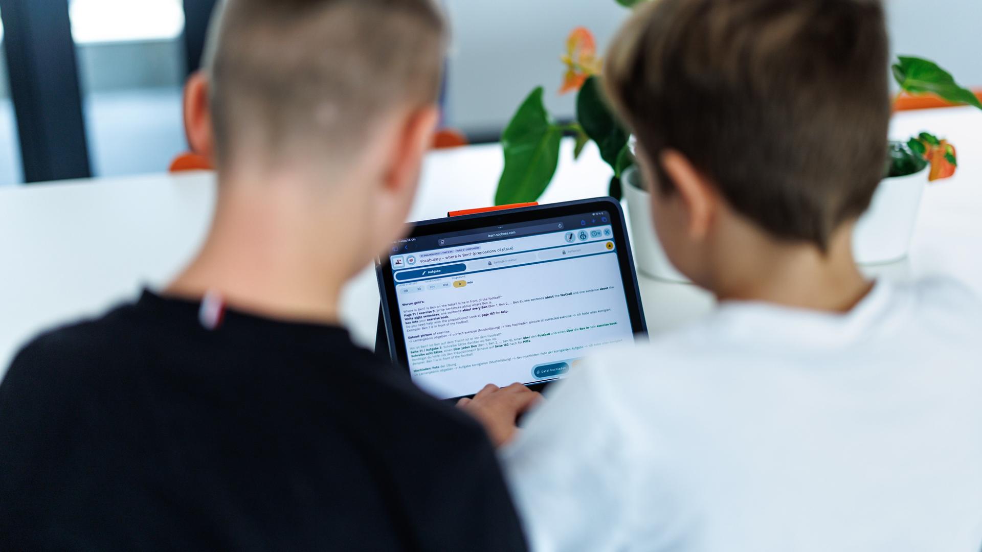 Zwei Kinder sitzen in ihrem Klassenzimmer am Tablet und schauen gemeinsam darauf. Sie sind von hinten zu sehen und damit nicht zu erkennen.