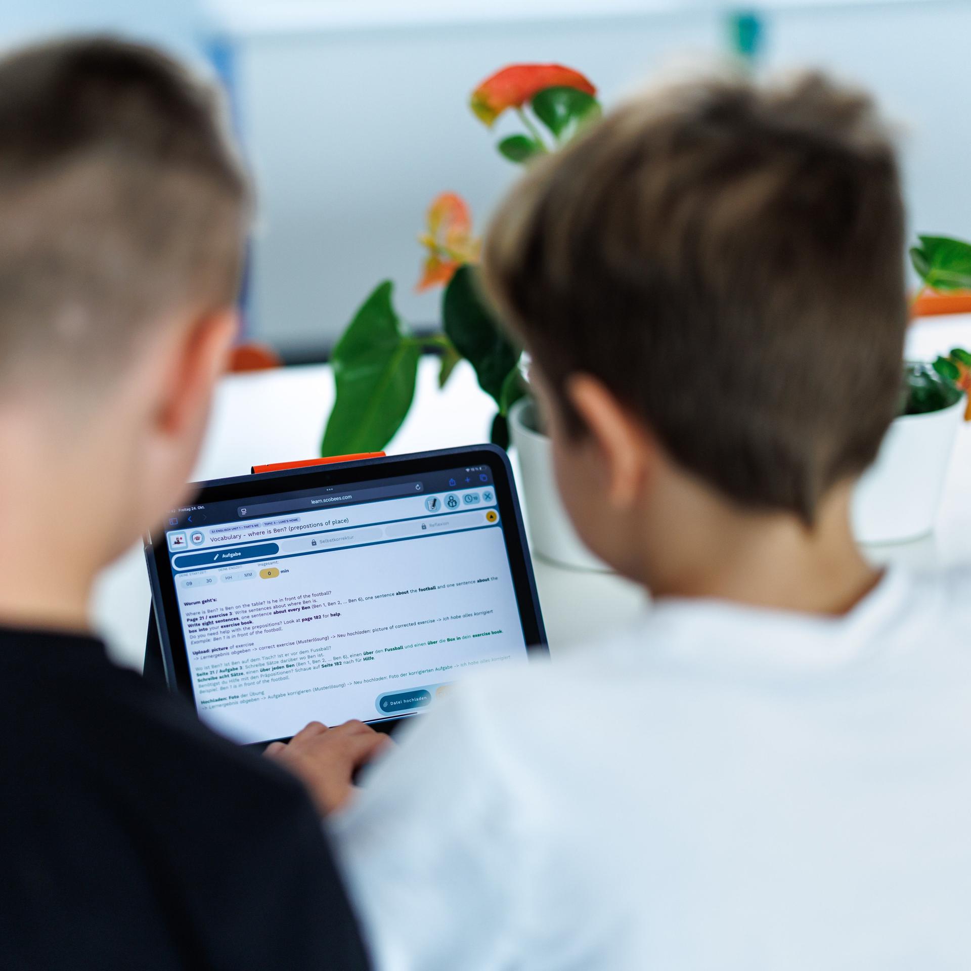 Zwei Kinder sitzen in ihrem Klassenzimmer am Tablet und schauen gemeinsam darauf. Sie sind von hinten zu sehen und damit nicht zu erkennen.