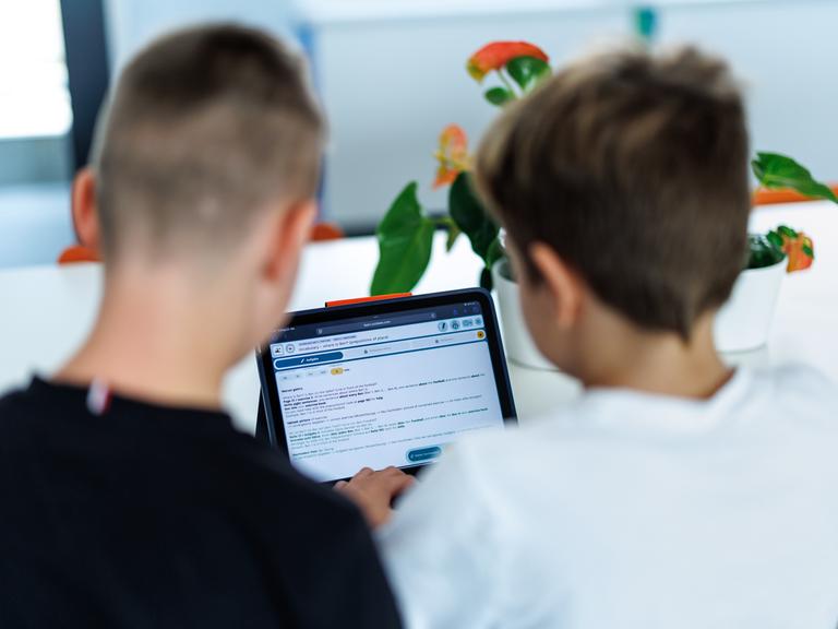 Zwei Kinder sitzen in ihrem Klassenzimmer am Tablet und schauen gemeinsam darauf. Sie sind von hinten zu sehen und damit nicht zu erkennen. Zwei Kinder sitzen in ihrem Klassenzimmer am Tablet und schauen gemeinsam darauf. Sie sind von hinten zu sehen und damit nicht zu erkennen.