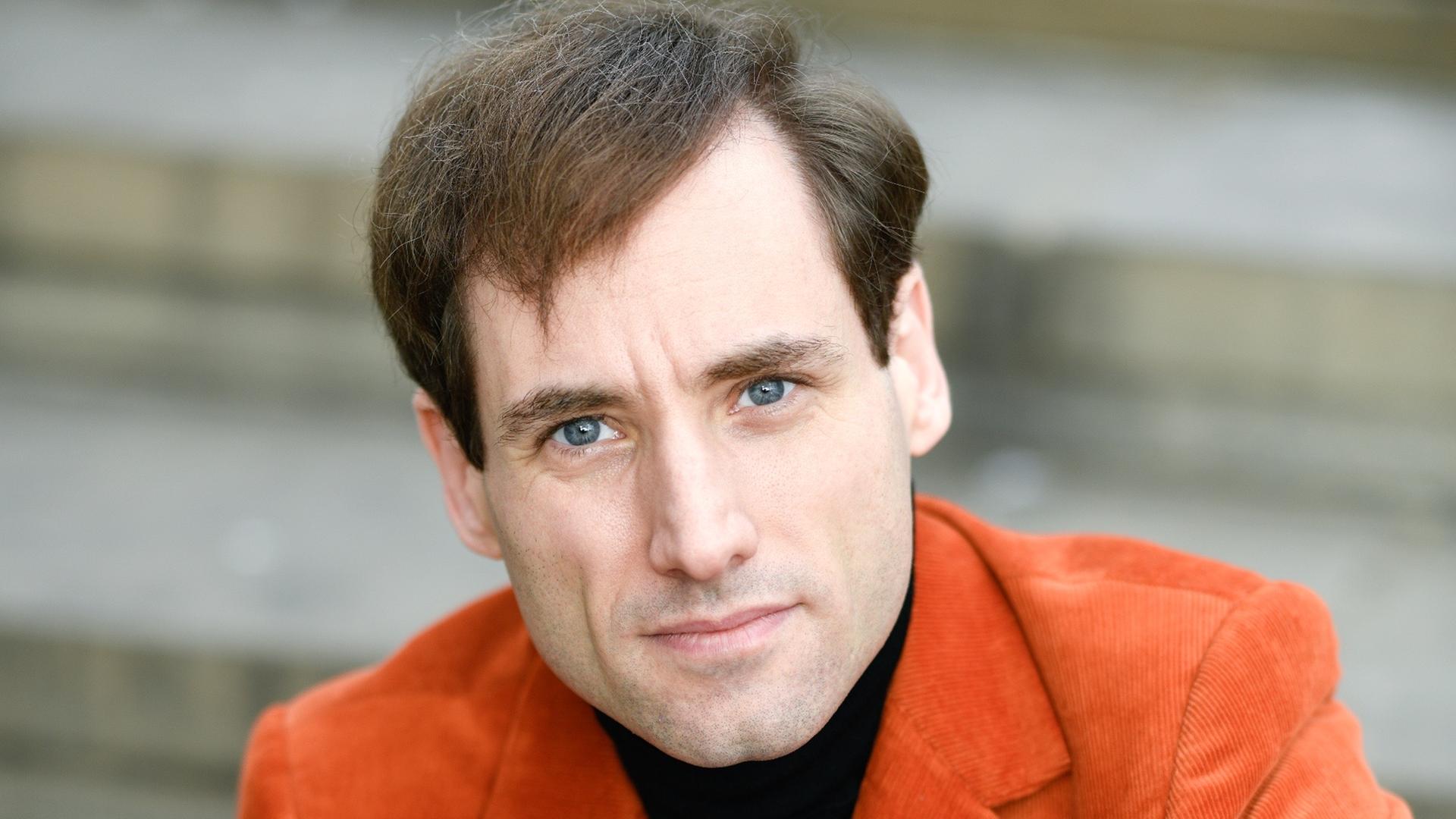 Ein Mann mit braunen Haaren und blauen Augen sitzt auf einer Treppe. Er schaut direkt in die Kamera. Er trägt ein orangefarbenes Cordjackett über einem schwarzen Shirt. Ein Mann mit braunen Haaren und blauen Augen sitzt auf einer Treppe. Er schaut direkt in die Kamera. Er trägt ein orangefarbenes Cordjackett über einem schwarzen Shirt.