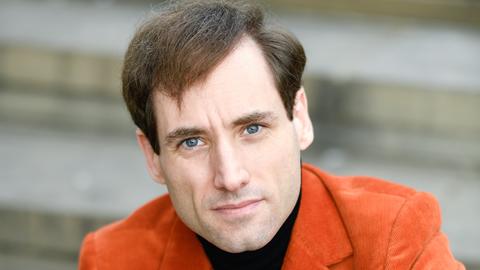 Ein Mann mit braunen Haaren und blauen Augen sitzt auf einer Treppe. Er schaut direkt in die Kamera. Er trägt ein orangefarbenes Cordjackett über einem schwarzen Shirt. 